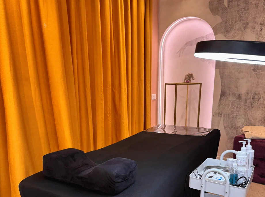 Cabinetul de electroepilare: pat de tratament negru, lampă cu lupă și draperie aurie, în fața peretelui roz